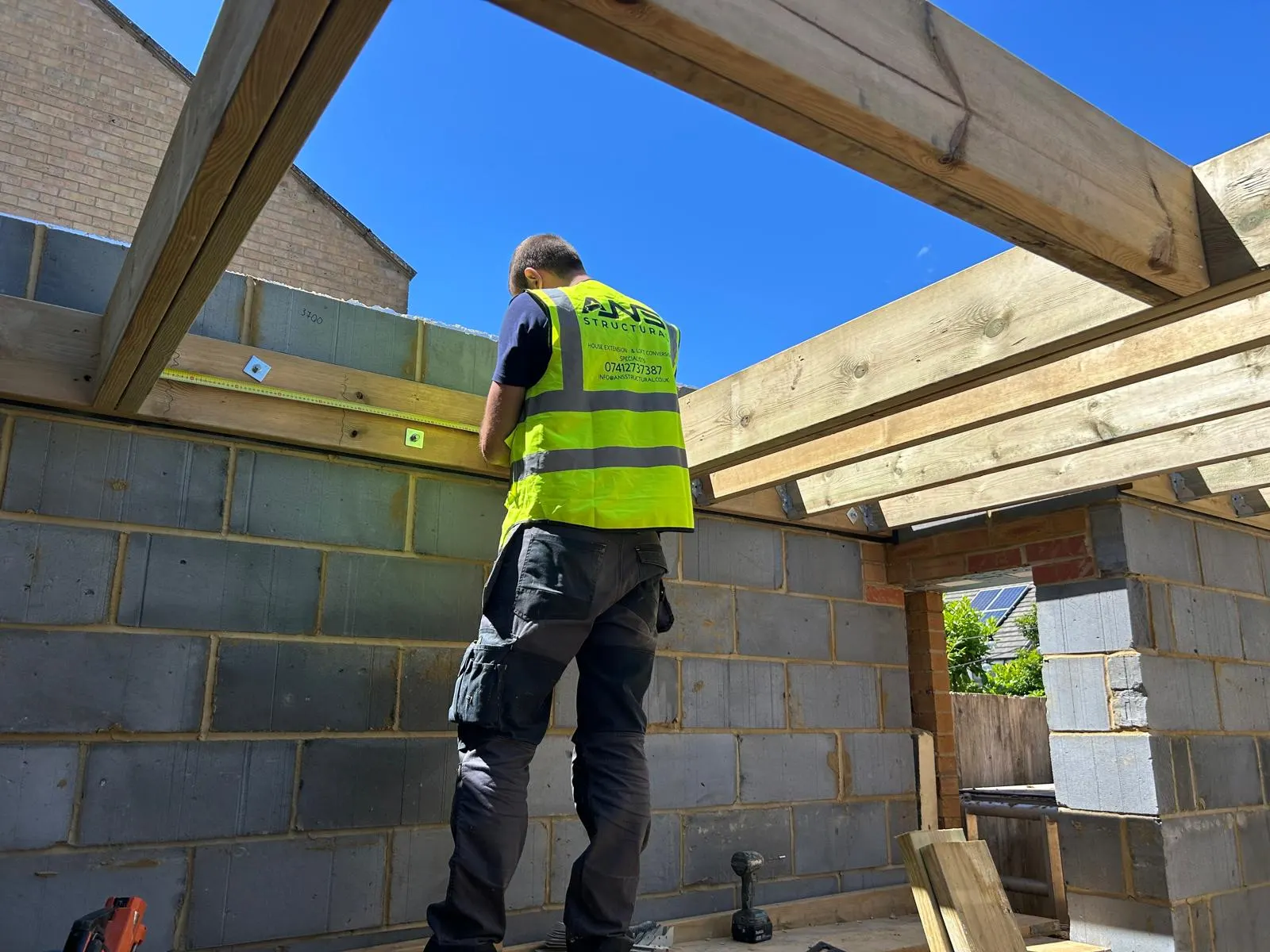 ANS Structural carpenter fixing new flat-roof joists on a house extension under a clear blue sky